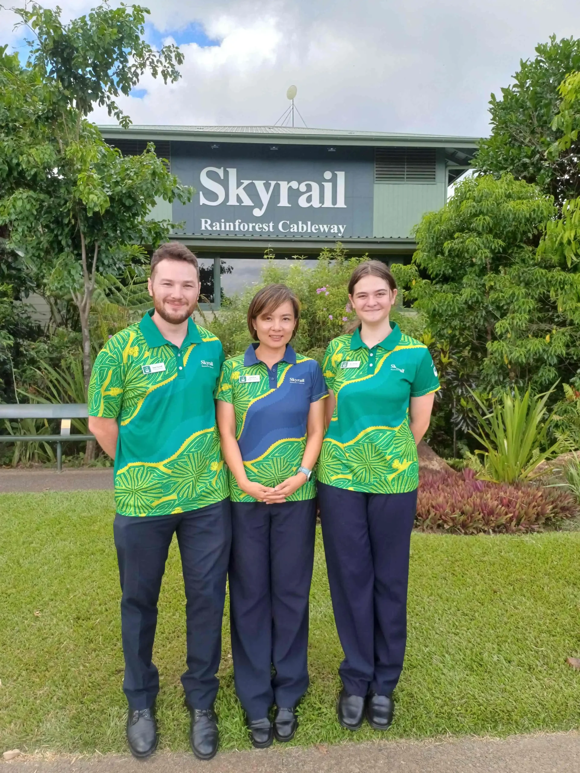 Two females and a male standing close together on a patch of grass in front of a tropical garden with a green building in the background. The name Skyrail Rainforest Cableway is in large letters as a logo on the building.