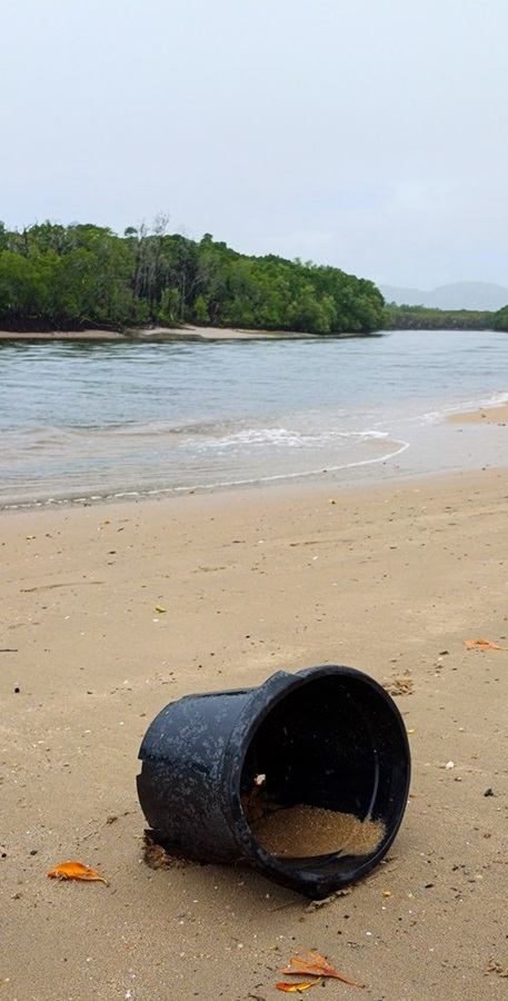 Large black rubbish bin partially filled with sand discarded on the beach