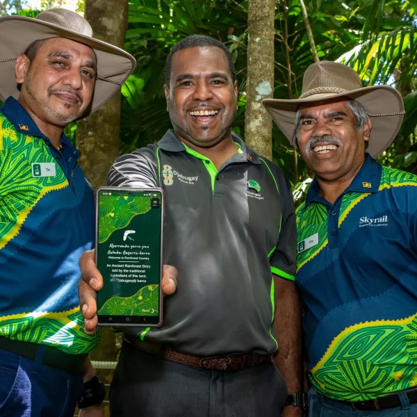 Three men stand together, smiling as the man in the middle holds out a phone into the foreground