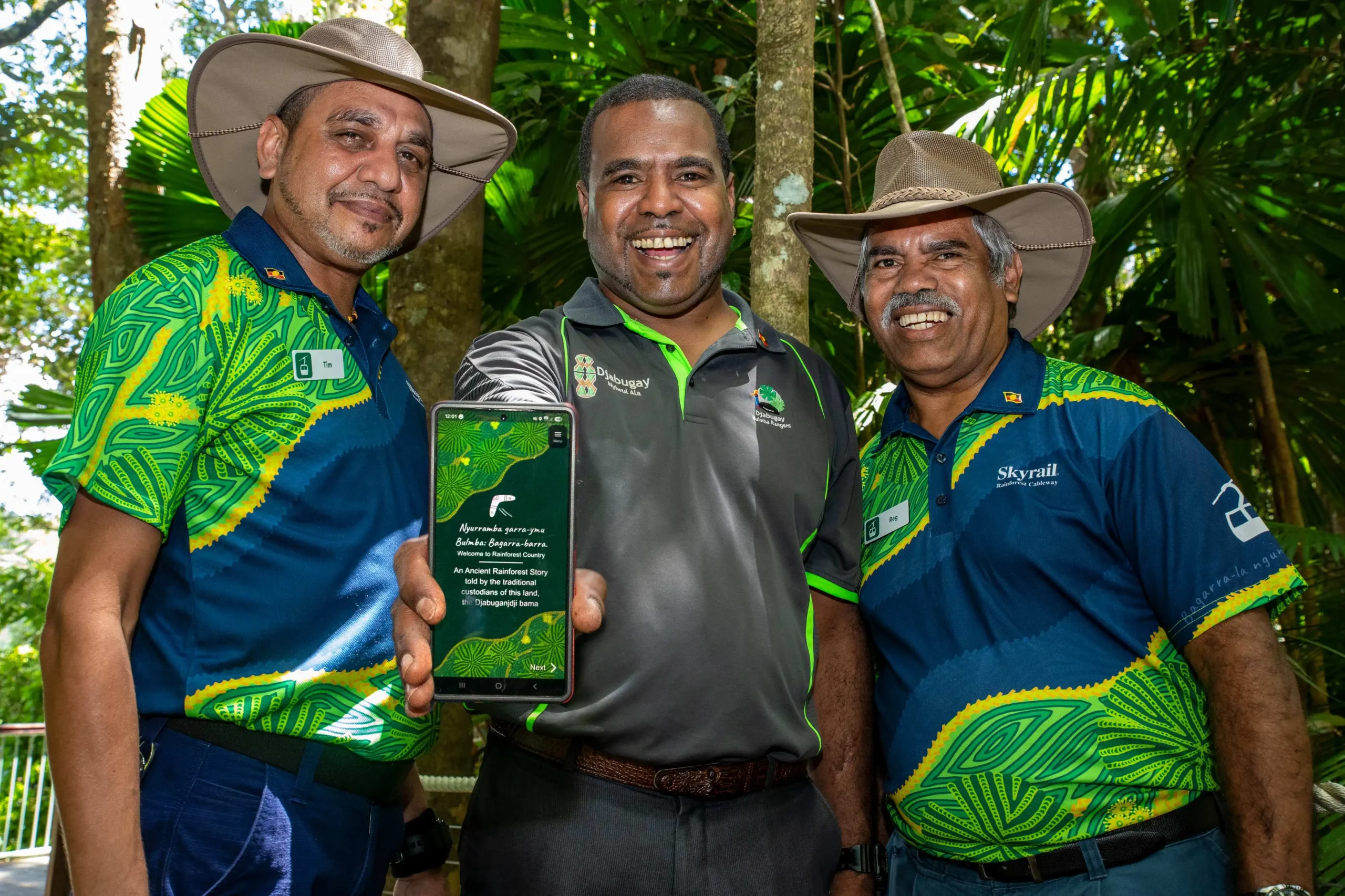 Three men stand together, smiling as the man in the middle holds out a phone into the foreground 