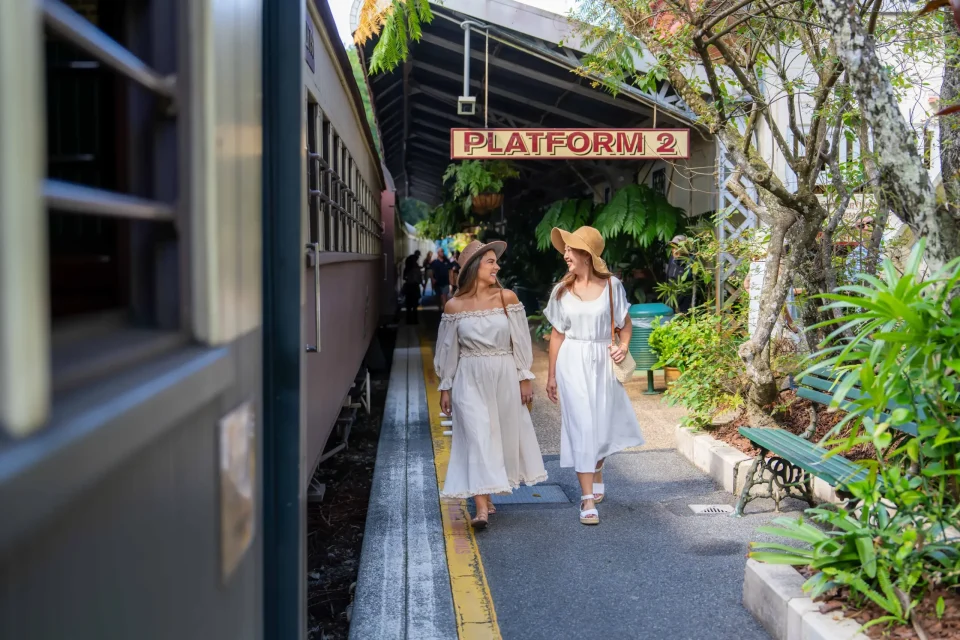 Two women in light coloured dresses walk alongside the train with the Kuranda station sign above them