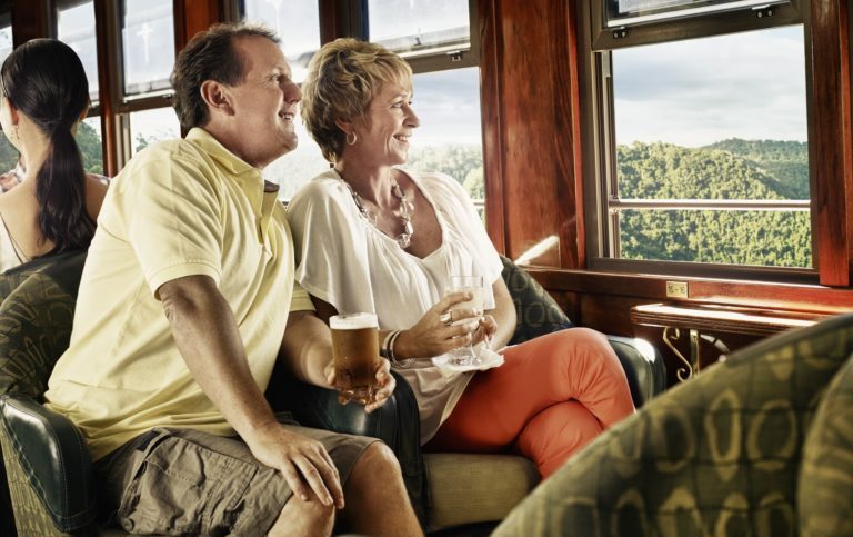 Couple sit close to an open window on the Kuranda train, having a drink and admiring the view across the Barron Gorge