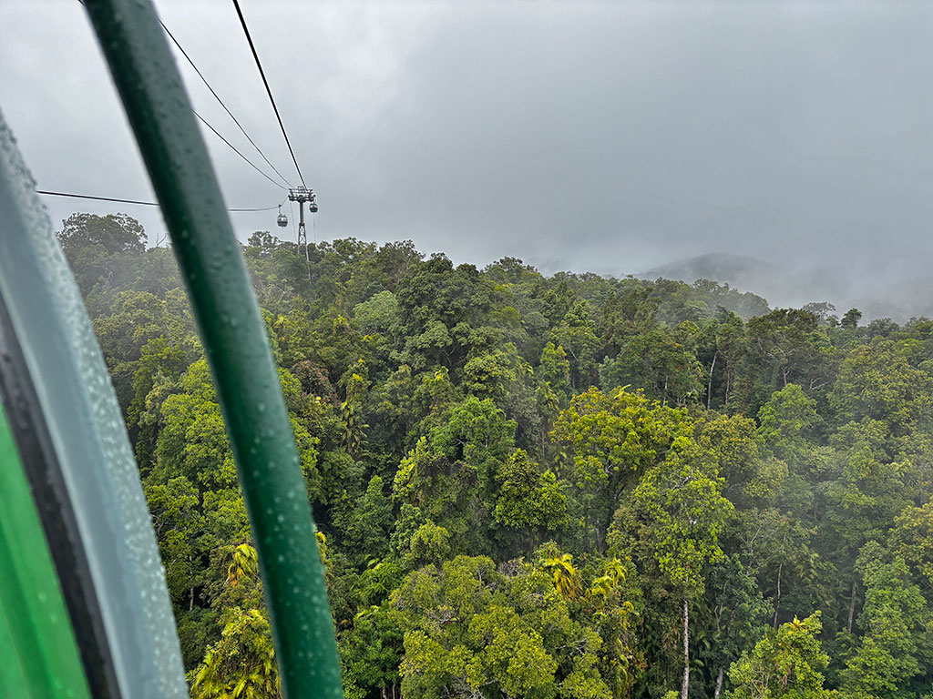 Looking out of a skyrail gondola window, back towards the cableway with more gondolas in the distance. The sky is darky and rainy with the mountains covered in mist