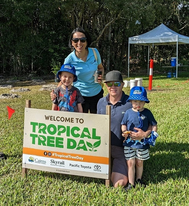 A family with two adults and two young children stand next to a large sign in a park reading welcome to tropical tree day