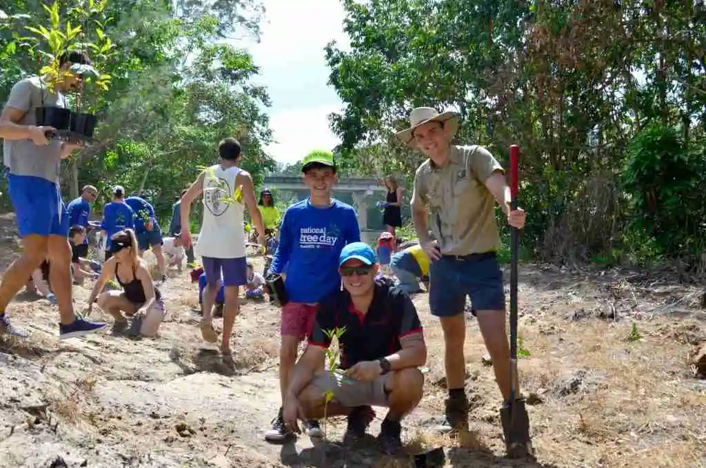 A man crouches on the ground with a fresh tree planted. A Ranger and smal boy stand behind him, all smiling at the camera