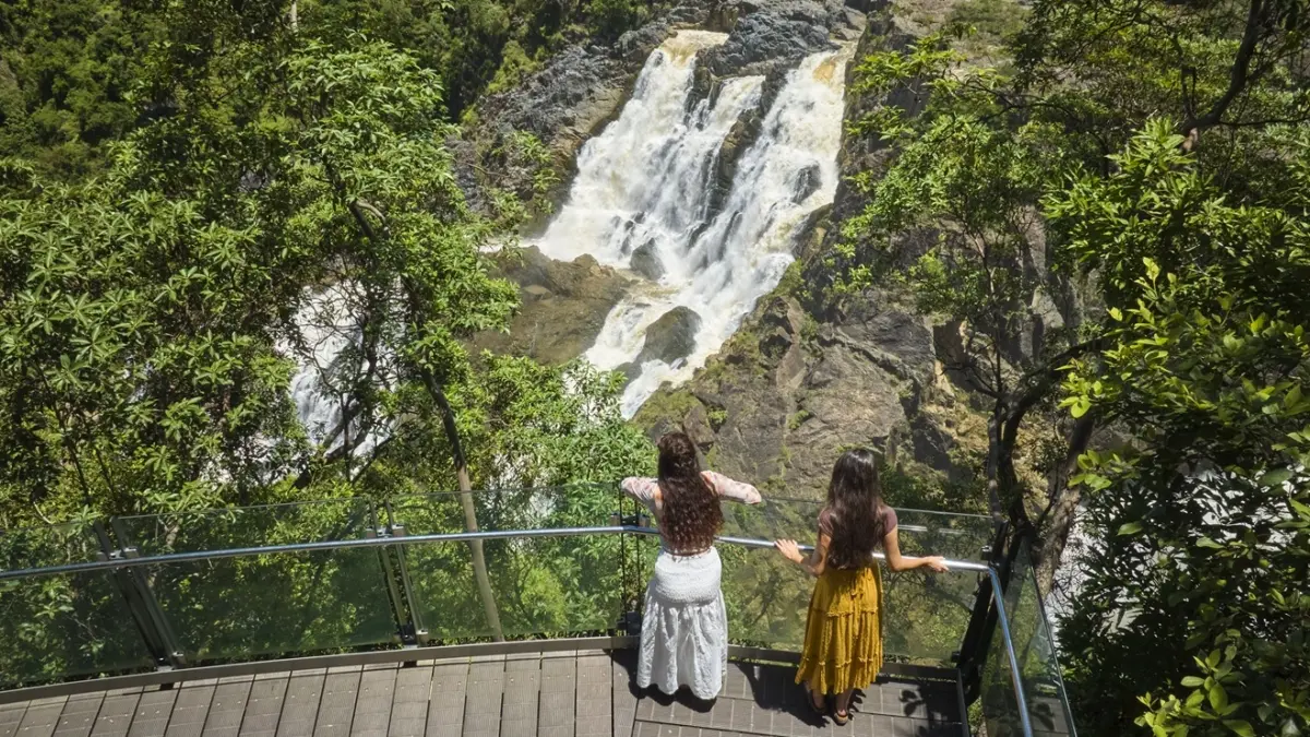 two girls in long dresses stand with their back to the camera, leaning against the glass railing of the edge lookout and looking towards Barron Falls