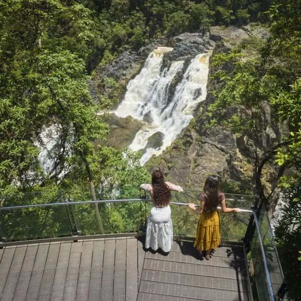 two girls in long dresses stand with their back to the camera, leaning against the glass railing of the edge lookout and looking towards Barron Falls